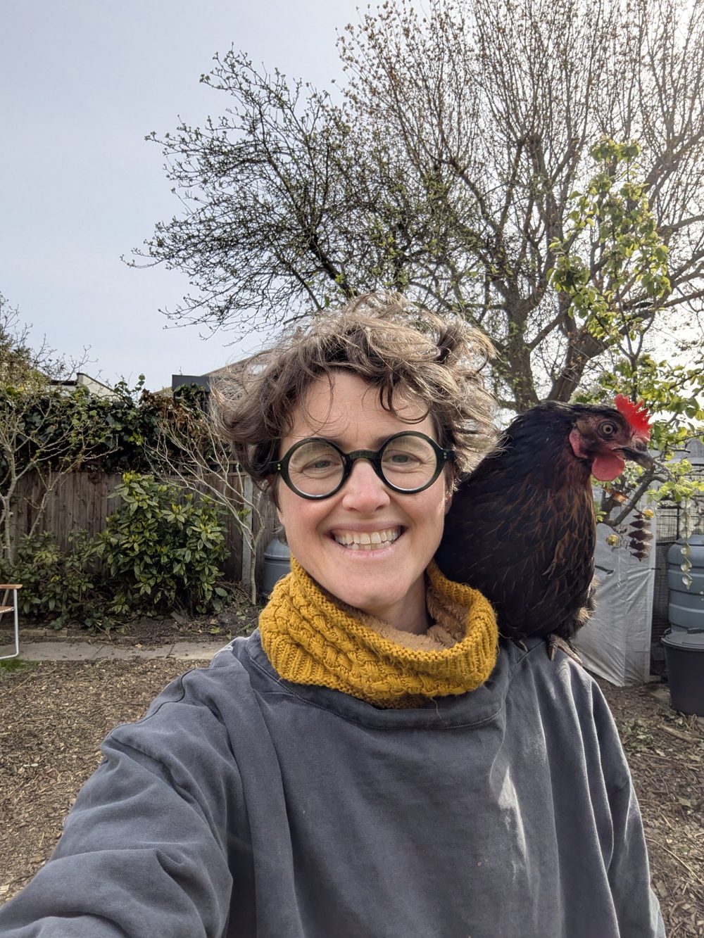 Holly smiling in her garden with a hen perched on her shoulder
